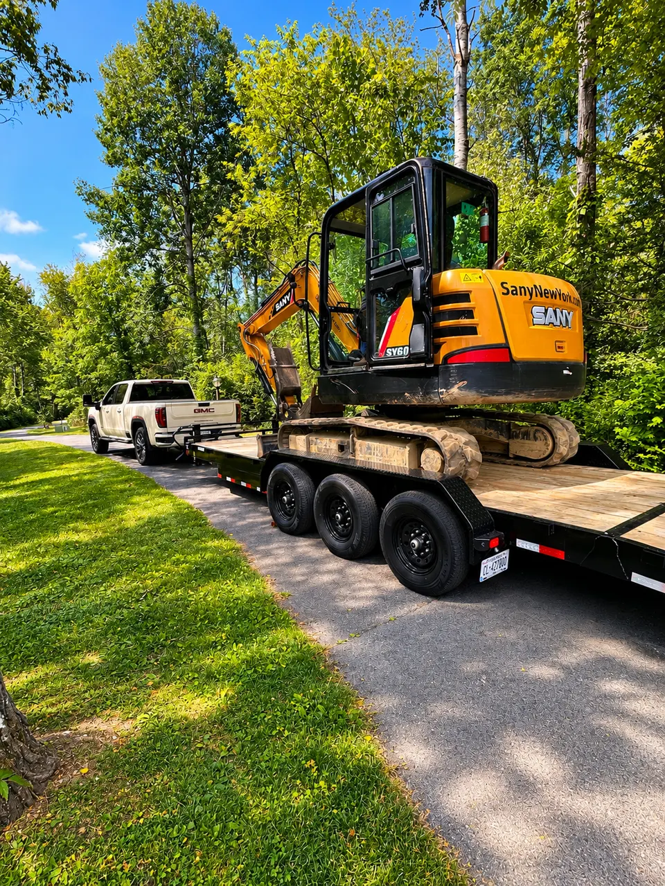 Mini excavator arriving on a trailer behind a white GMC pickup at a residential job site